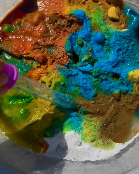 A pipette dripping color into fizzing baking soda in a glass bowl, in blue, orange and green.
