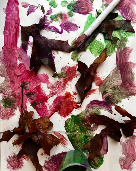 Autumn leaves pressed into pink, purple and green paint on paper, with a brush and a cup.