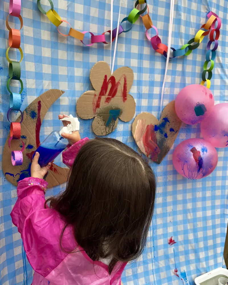 A child in a pink smock sprays blue paint onto cardboard shapes taped to a gingham backdrop.